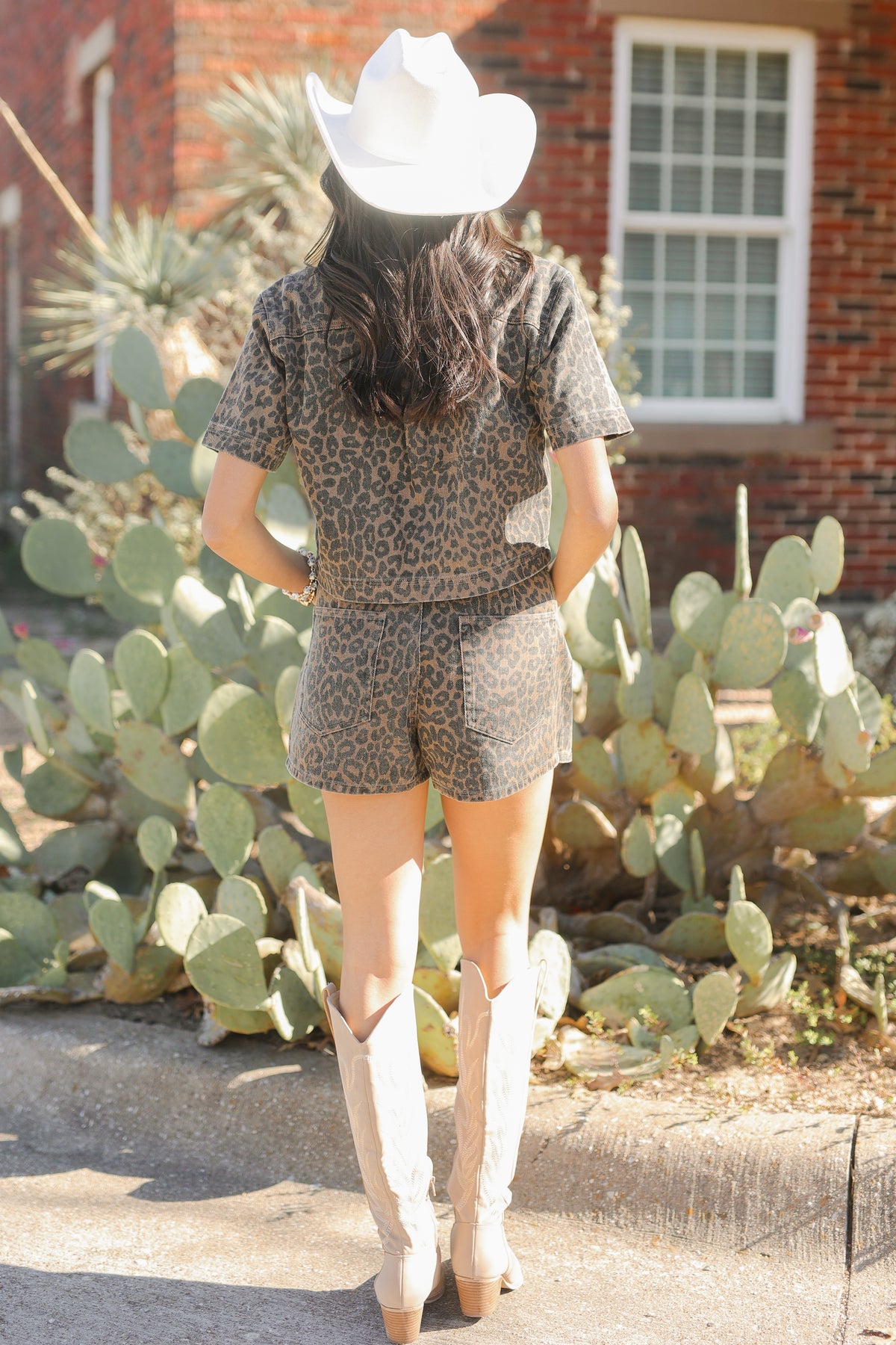 Person wearing a leopard print outfit with a cowboy hat and boots in front of cacti.