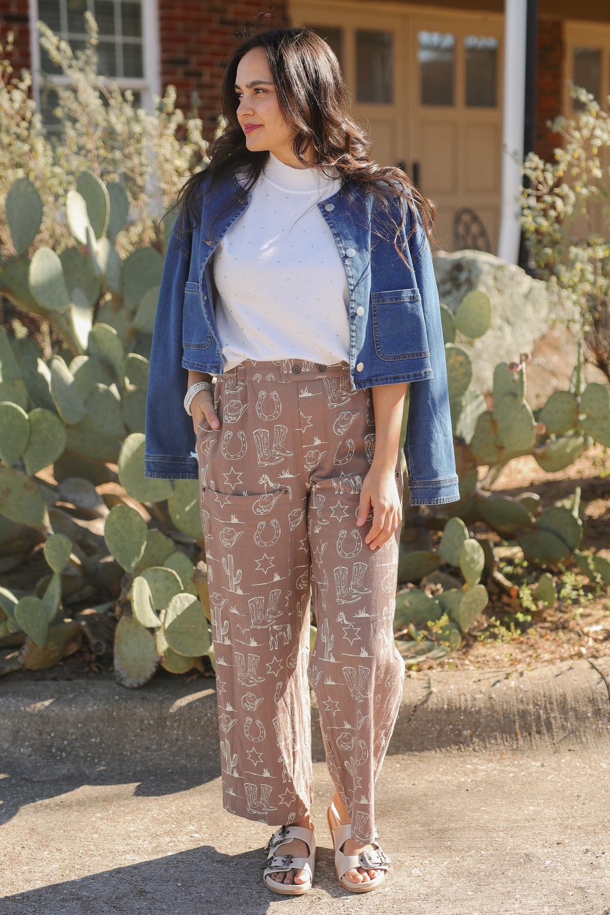 Woman wearing a denim jacket, white shirt, and patterned pants standing in front of cacti.