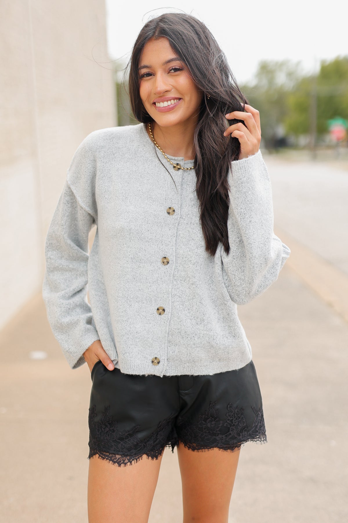 Brunette model standing outside wearing the Ashford Everyday Knit Cardigan in Heather Gray featuring a soft textured knit, round neckline, tortoiseshell-style buttons, and relaxed fit, paired with black lace-trim shorts and the Kevin cowboy boots in black. Southern House Clothing