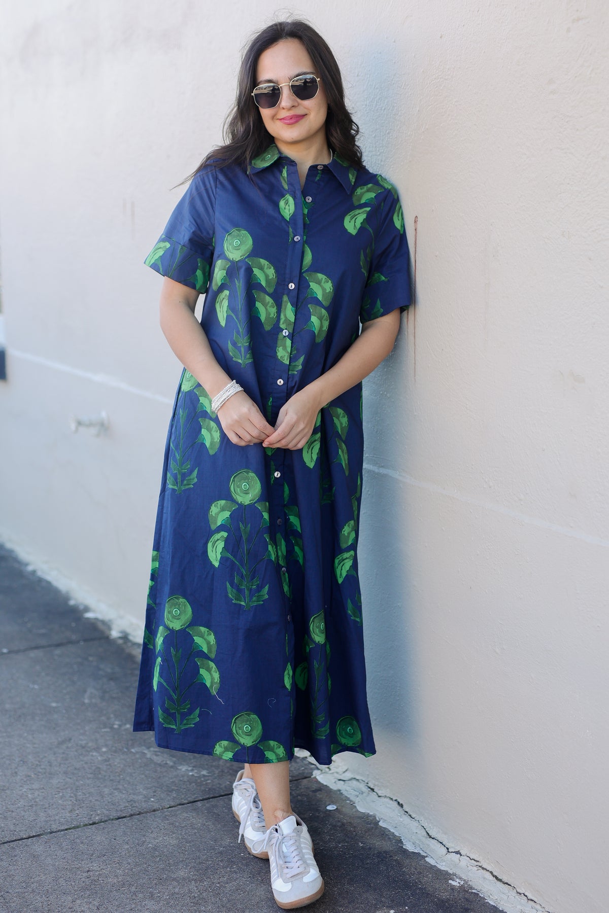 Woman wearing a long blue dress with green patterns against a white wall.
