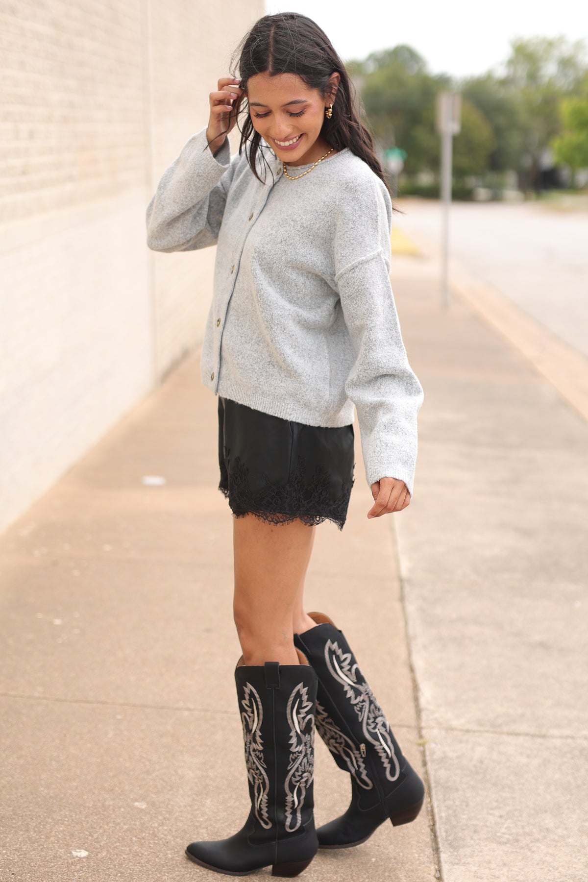 Brunette model standing outside wearing the Ashford Everyday Knit Cardigan in Heather Gray featuring a soft textured knit, round neckline, tortoiseshell-style buttons, and relaxed fit, paired with black lace-trim shorts and the Kevin cowboy boots in black. Southern House Clothing