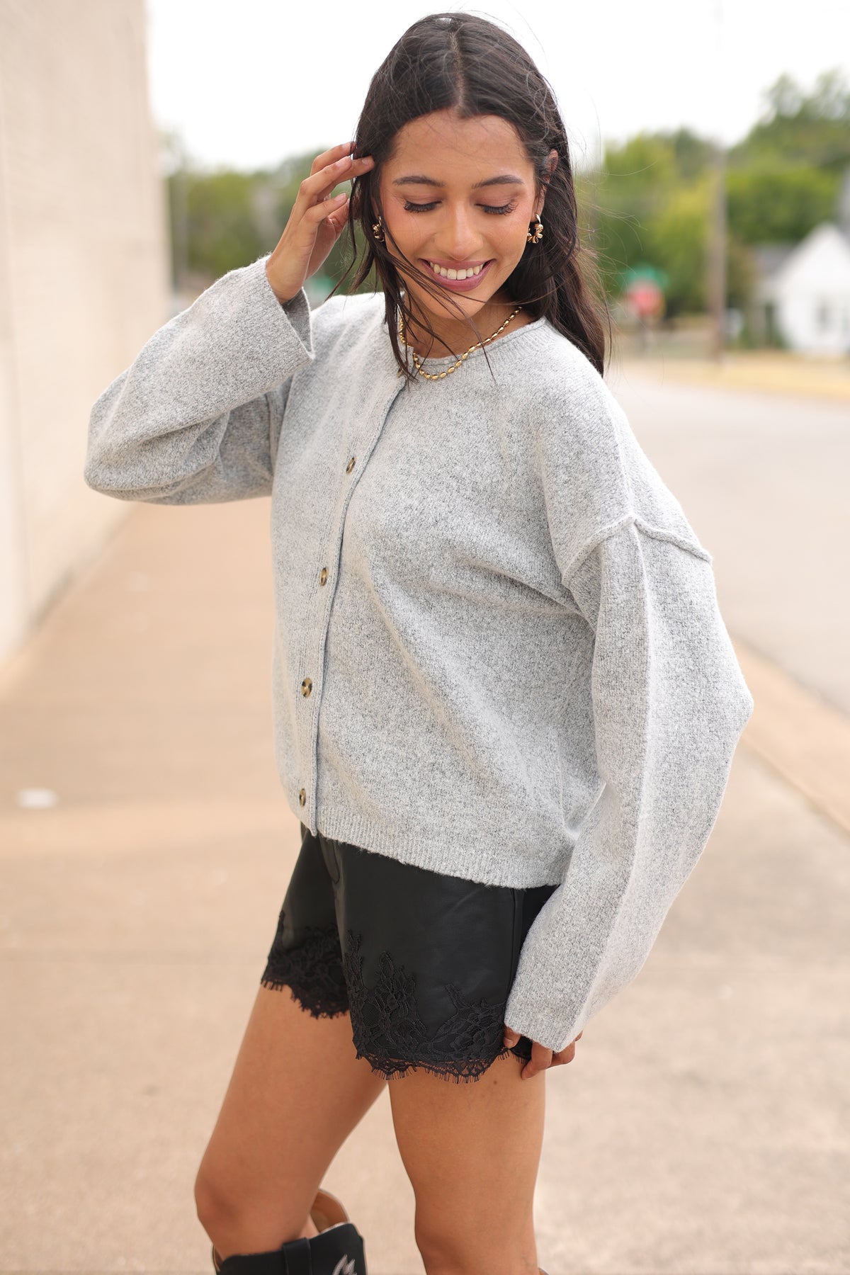 Brunette model standing outside wearing the Ashford Everyday Knit Cardigan in Heather Gray featuring a soft textured knit, round neckline, tortoiseshell-style buttons, and relaxed fit, paired with black lace-trim shorts and the Kevin cowboy boots in black. Southern House Clothing