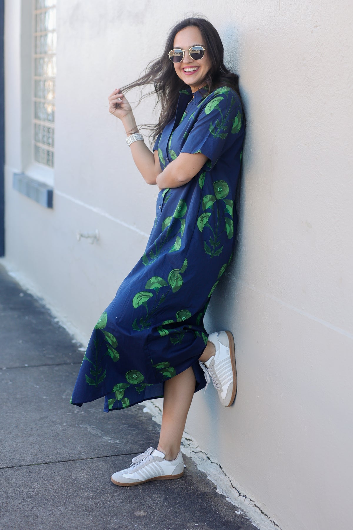 Woman wearing a blue dress with green leaf pattern leaning against a white wall.