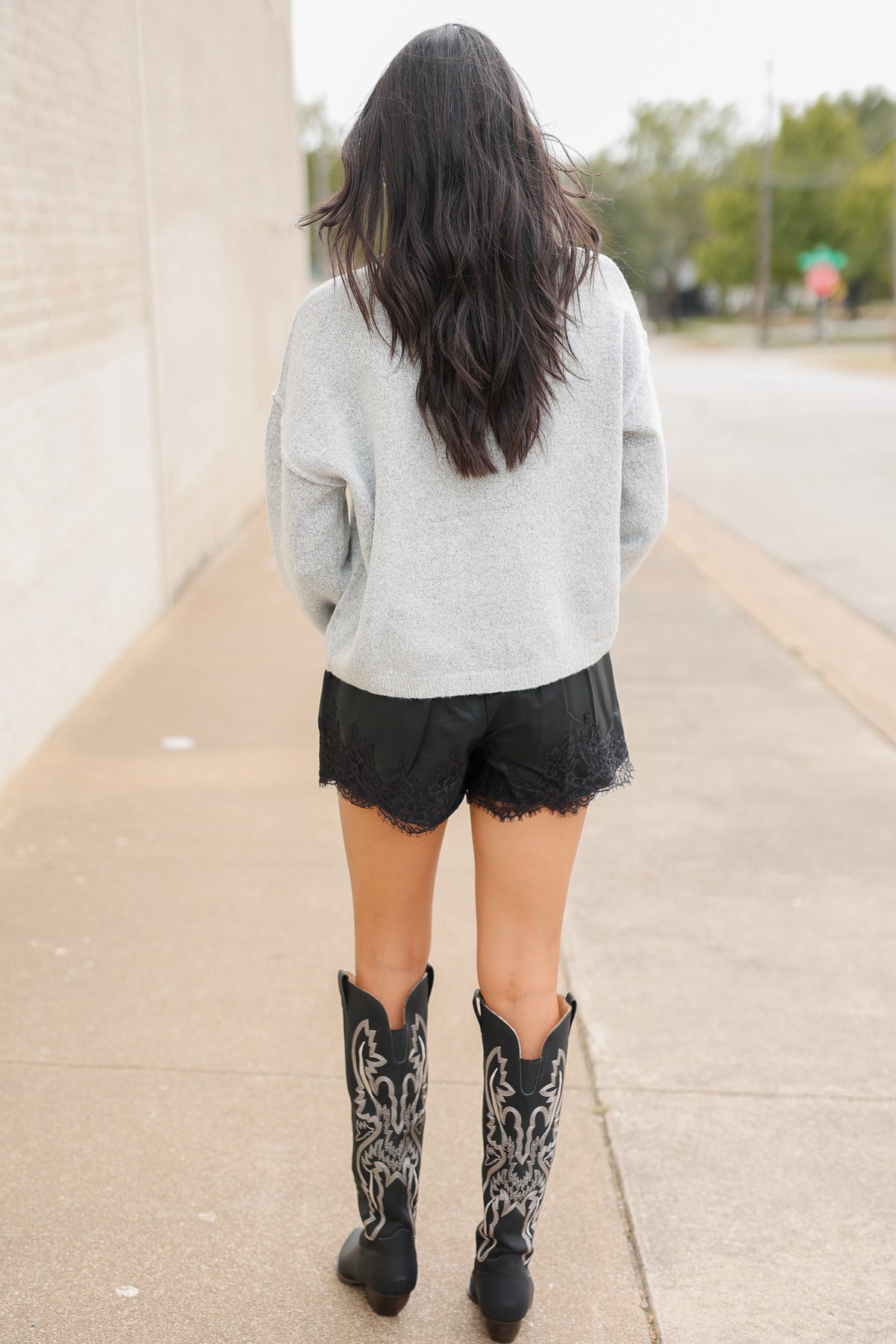 Brunette model standing outside wearing the Ashford Everyday Knit Cardigan in Heather Gray featuring a soft textured knit, round neckline, tortoiseshell-style buttons, and relaxed fit, paired with black lace-trim shorts and the Kevin cowboy boots in black. Southern House Clothing