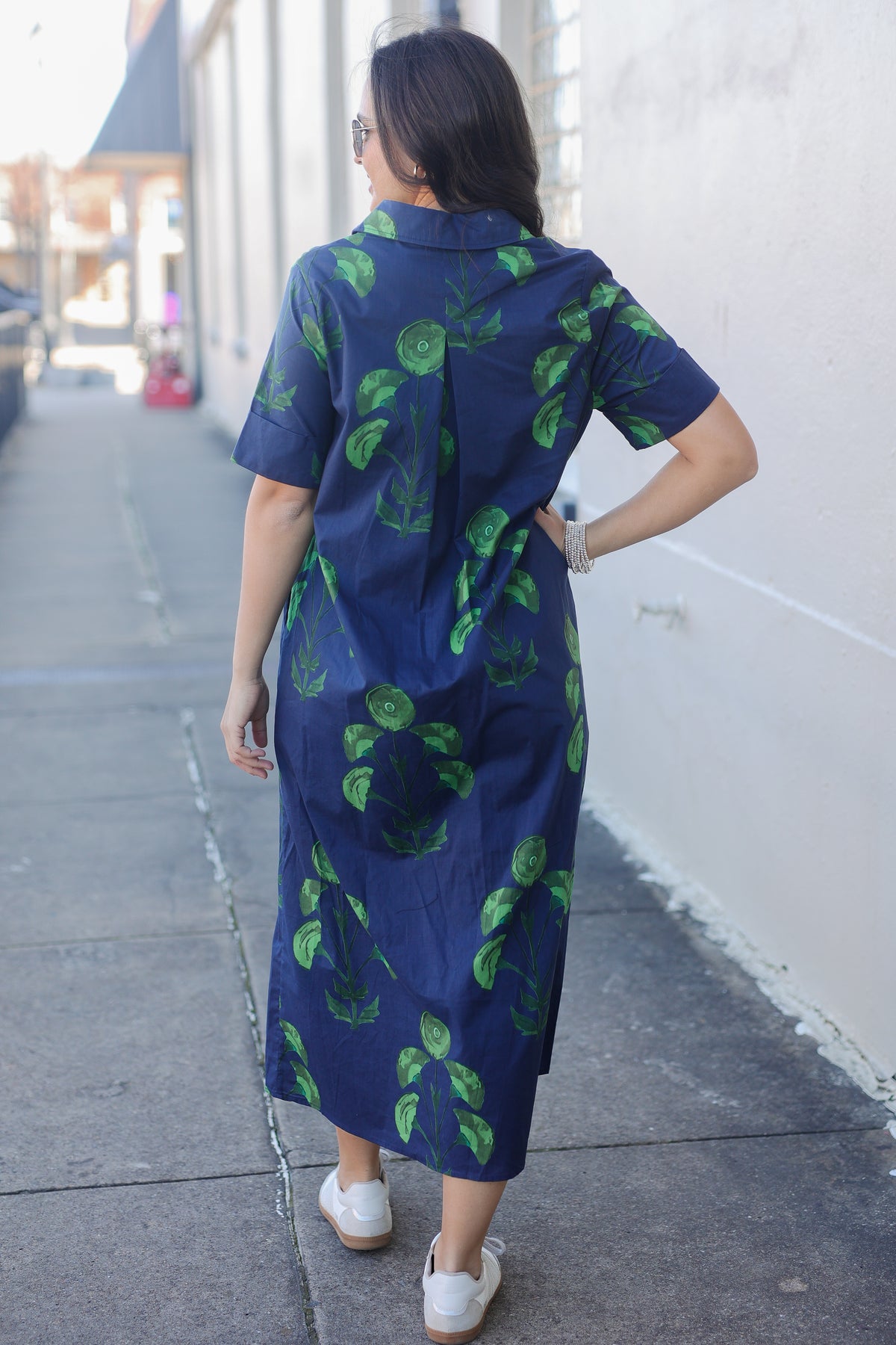 Woman wearing a blue dress with green leaf patterns walking on a sidewalk.