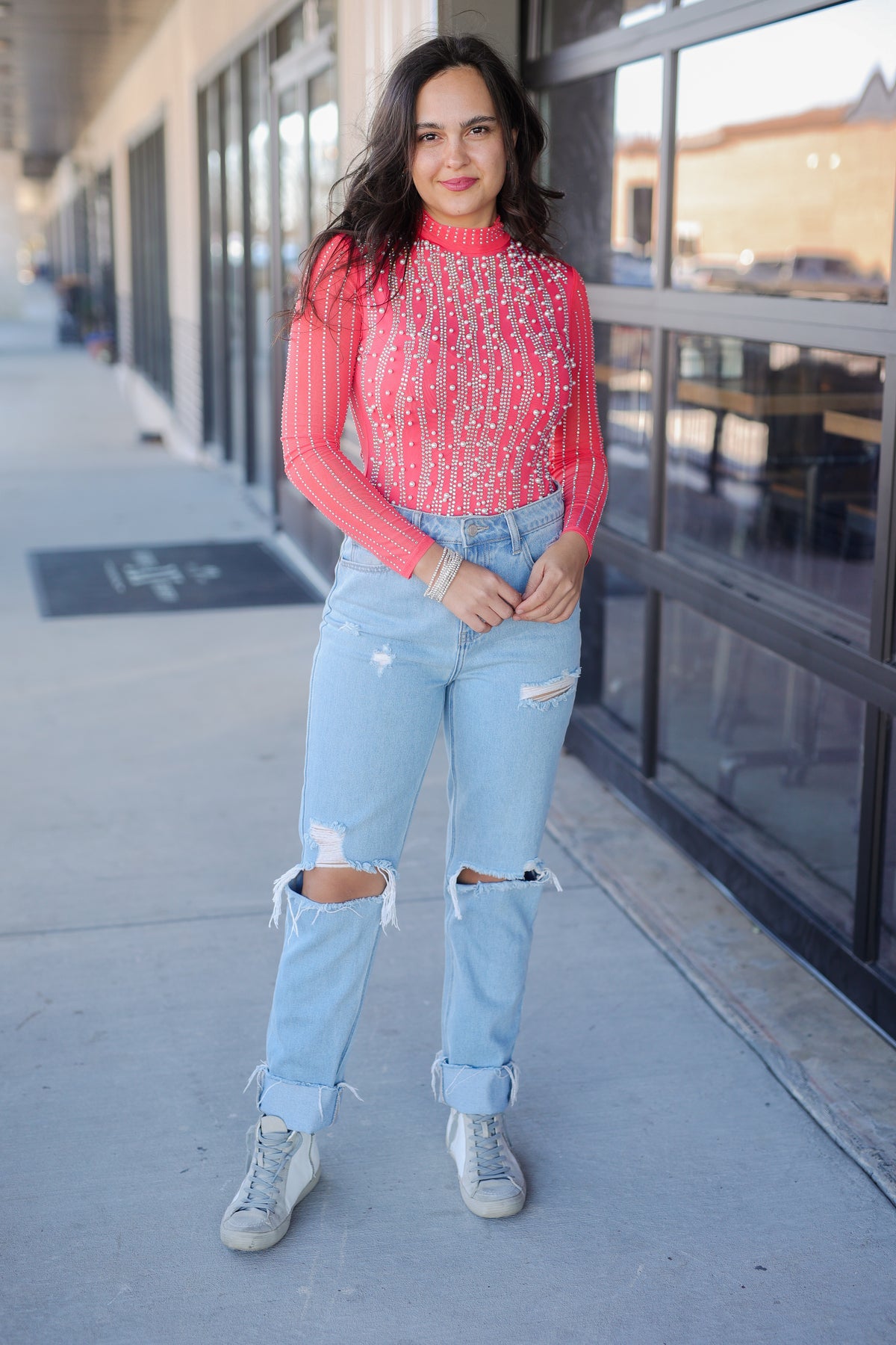 Woman wearing a red top and light blue jeans standing on a sidewalk.