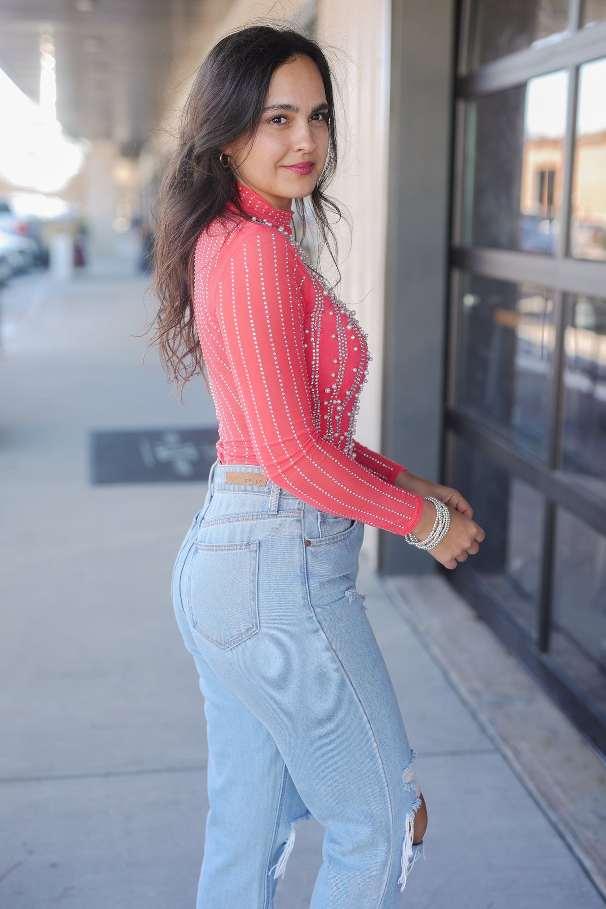 Woman wearing a red top and light blue jeans standing on a sidewalk.