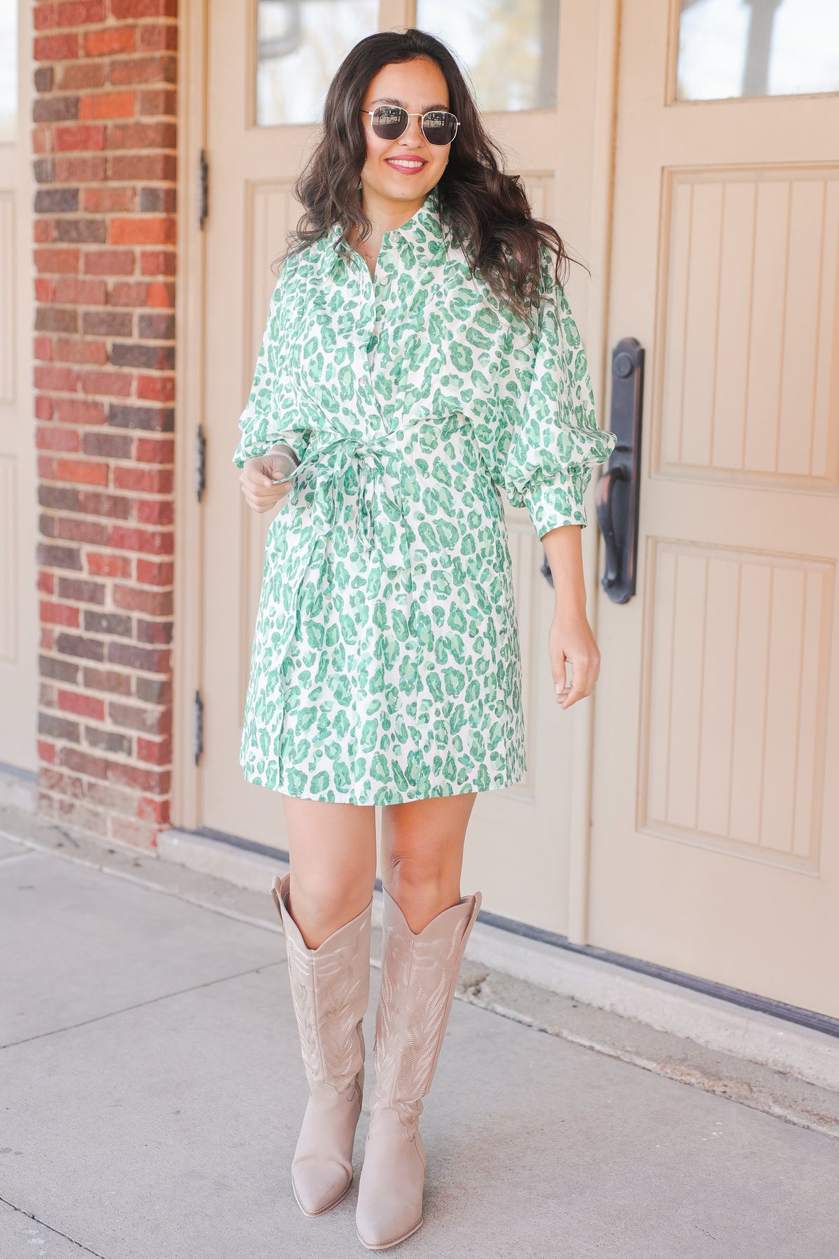 Woman wearing a green patterned dress and beige knee-high boots standing in front of a door.