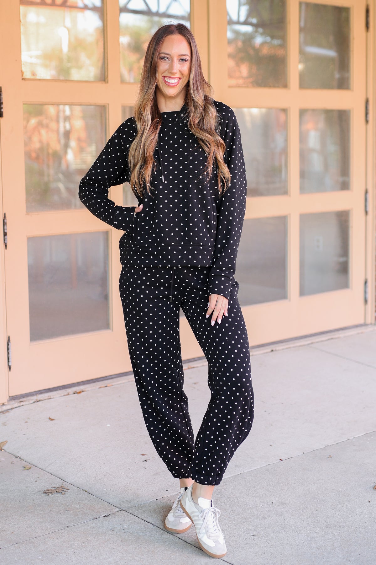 A woman modeling a matching black and white polka dot lounge set, consisting of a long-sleeve top and jogger pants, standing on a concrete sidewalk in front of a peach-colored glass door.