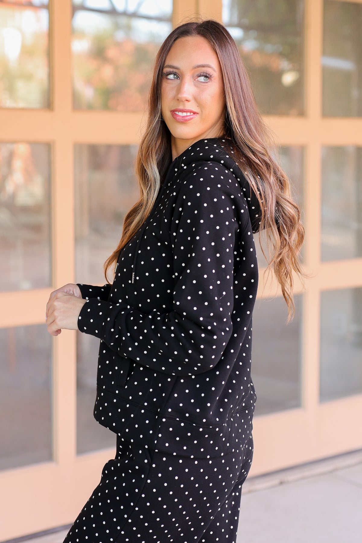A woman wearing a black long-sleeve hoodie and matching pants with white polka dots, standing in front of a beige glass-paneled building.
