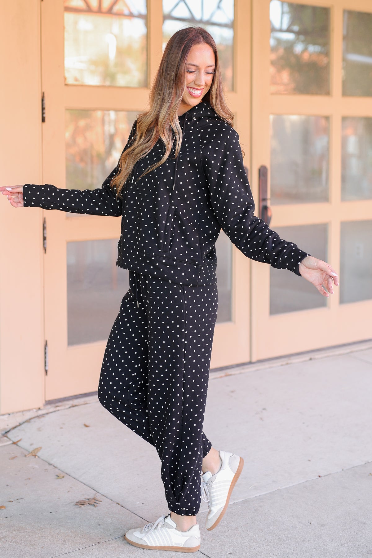 A woman modeling a matching black and white polka dot lounge set, featuring a long-sleeve top and jogger pants, paired with white sneakers, standing in front of peach-colored glass doors.