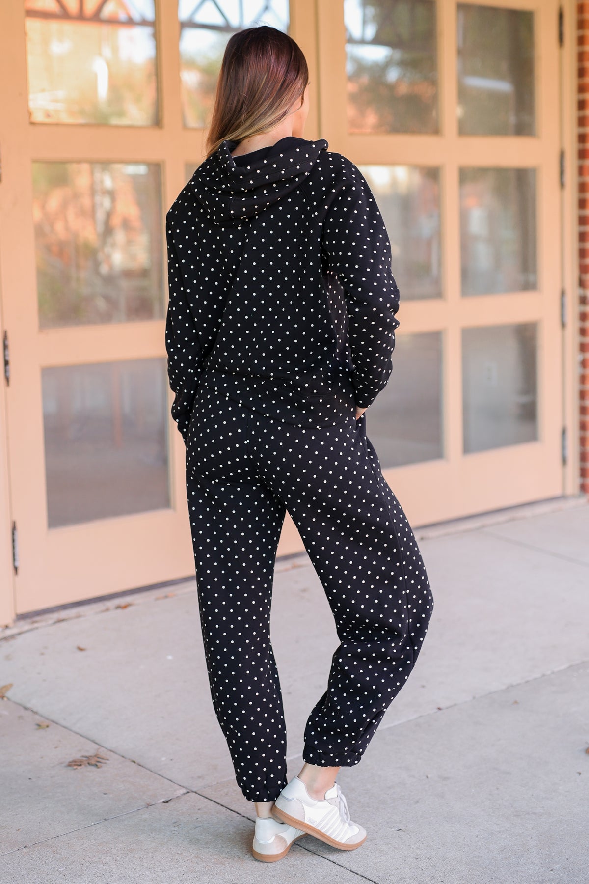 A woman wearing a black hooded sweatshirt and matching jogger pants with a white polka dot pattern, photographed from behind in front of a peach-colored glass door.