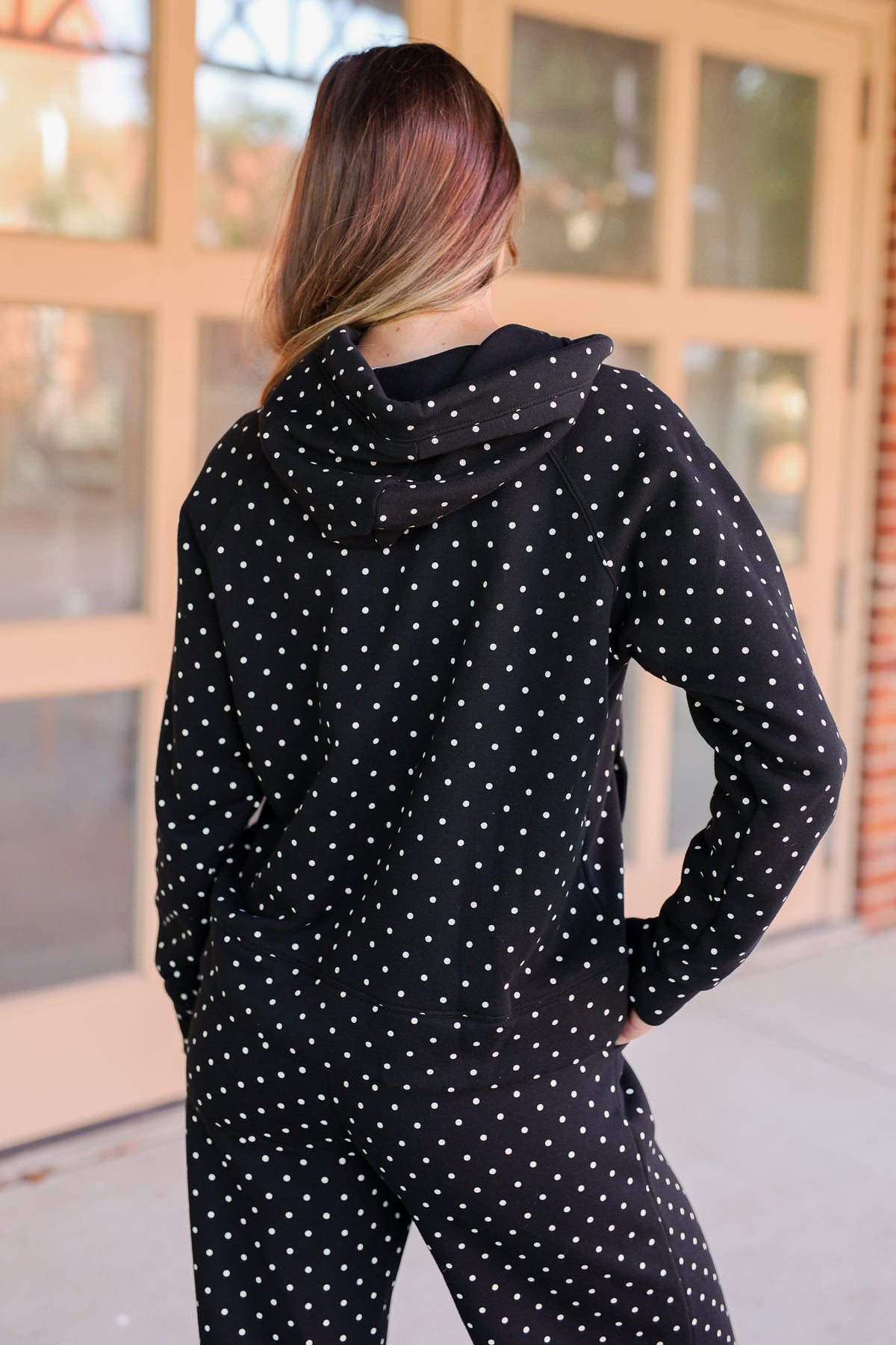 A woman viewed from behind wearing a black hooded jumpsuit with a white polka dot pattern, standing outdoors in front of a building with large windows.