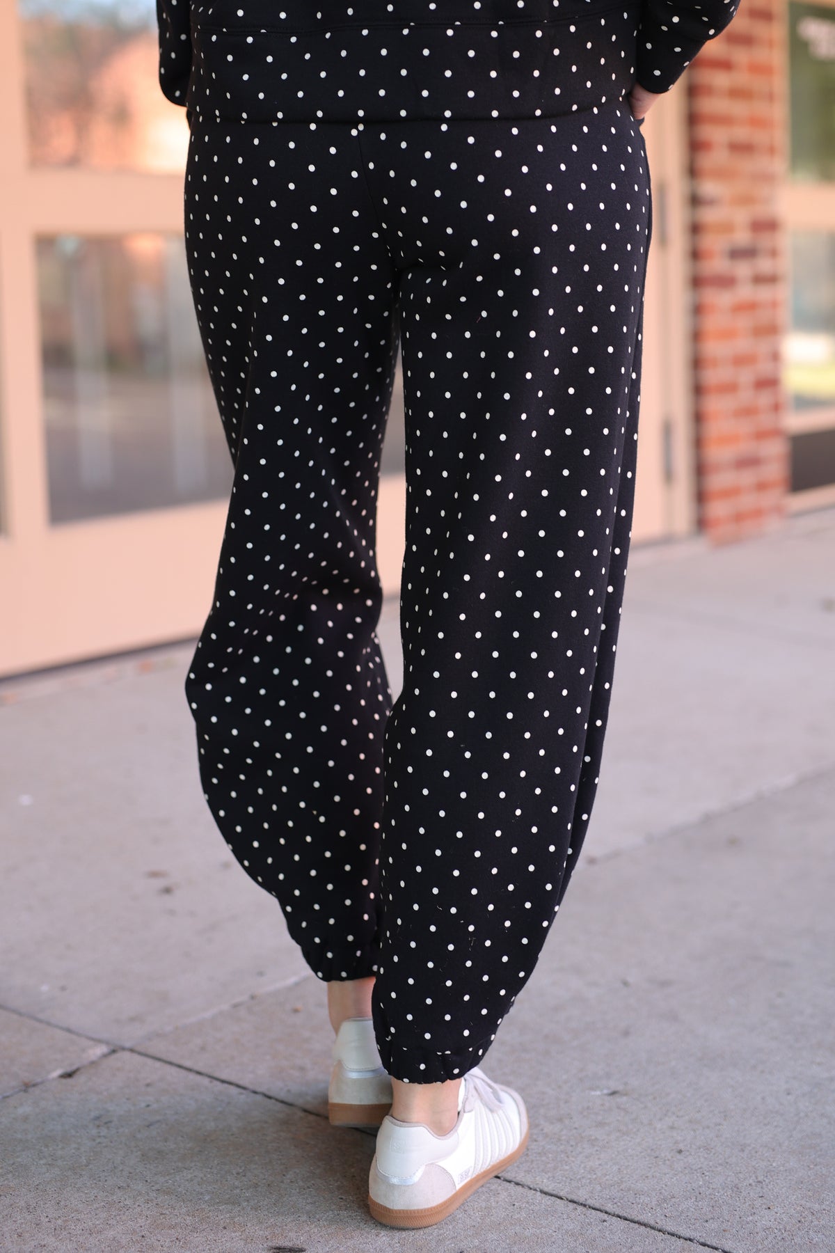 Black wide-leg pants with a white polka dot pattern, shown on a model from the back walking on a sidewalk.