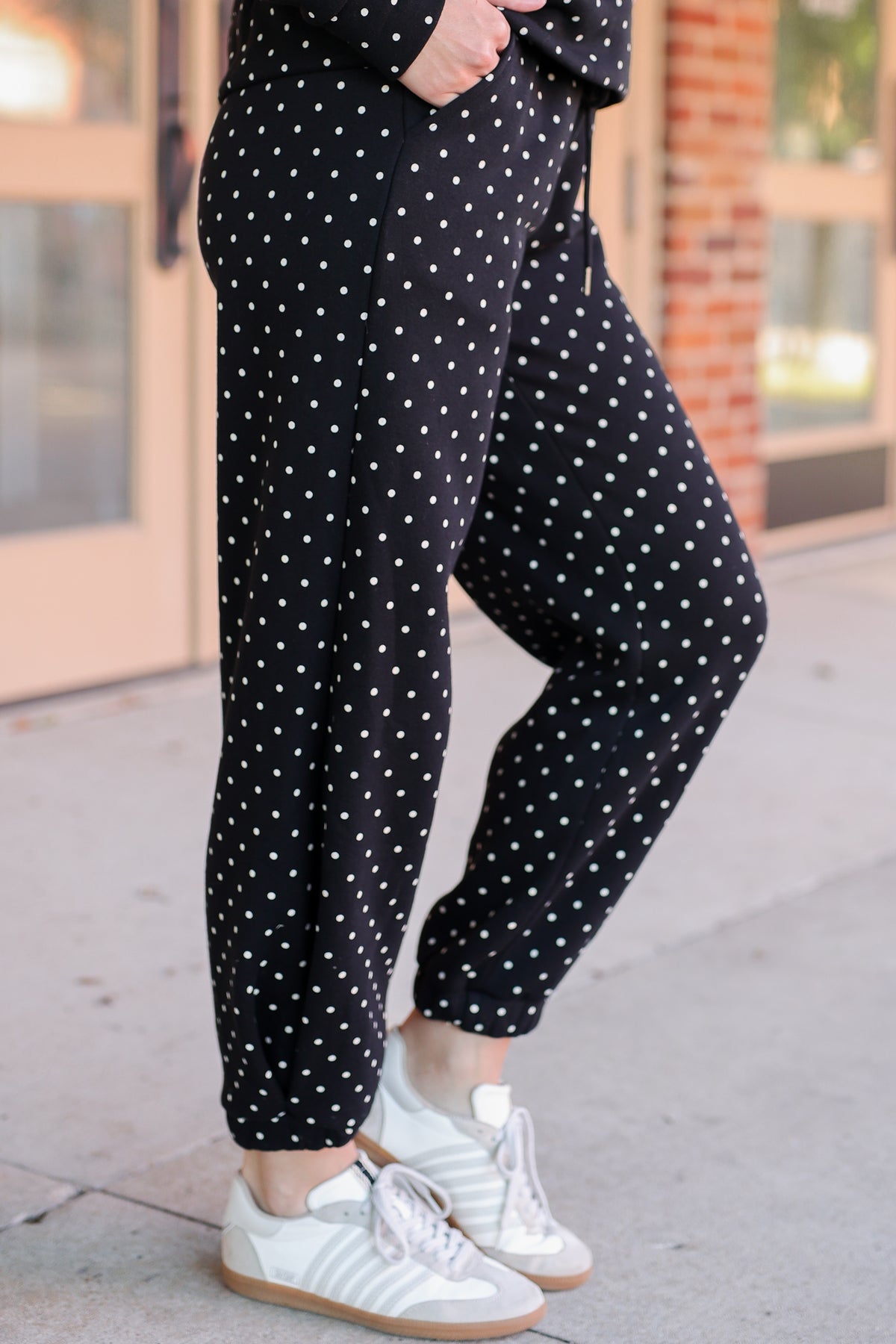 Black jogger pants with a white polka dot pattern, worn by a model standing on a sidewalk in front of a building.