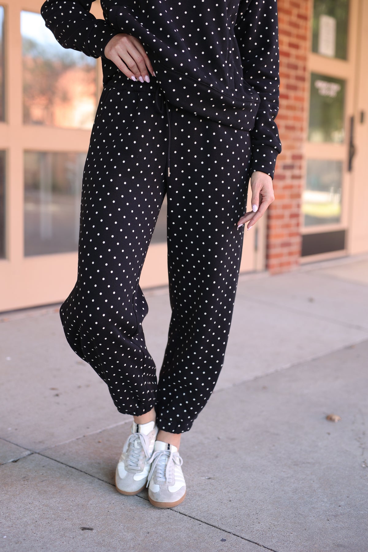 A person wearing a matching black and white polka dot lounge set, featuring a long-sleeve top and jogger-style pants, paired with white sneakers, standing on a sidewalk in front of a building.
