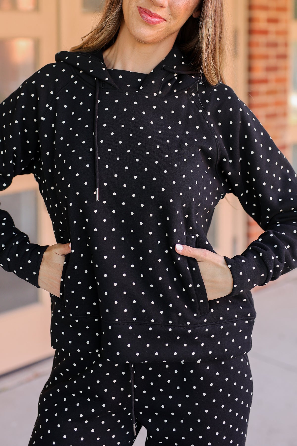 A woman wearing a black hooded sweatshirt and matching pants with a white polka dot pattern, standing with her hands in the pockets of her hoodie.