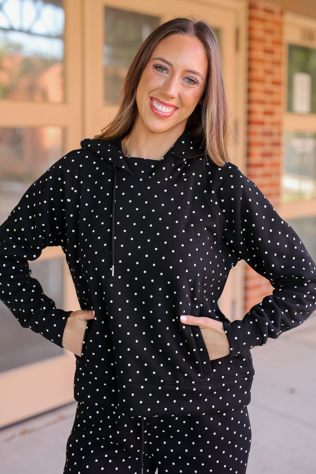 A woman smiling and posing in a black long-sleeve hoodie with white polka dots, set against a blurred storefront background.