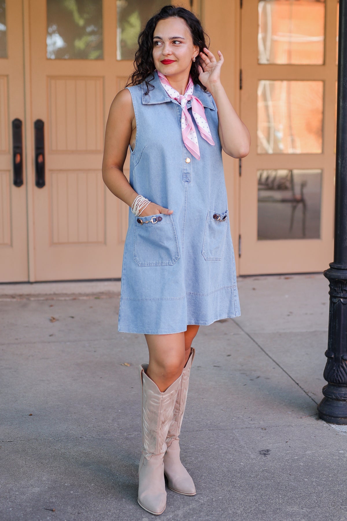 Woman in a denim dress with a pink scarf standing on a sidewalk.