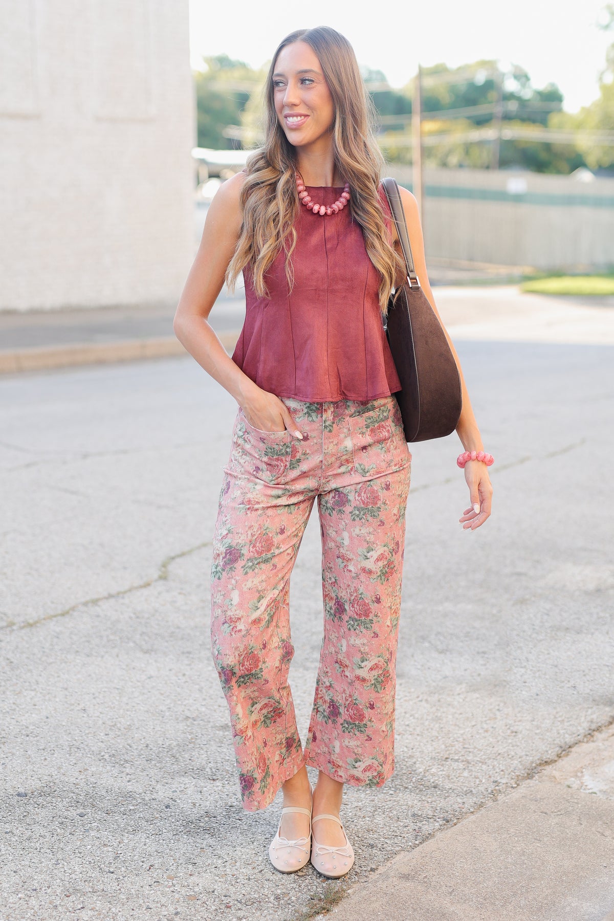 A woman wearing a sleeveless dusty rose top and floral pants, accessorized with a chunky pink beaded necklace and a brown shoulder bag, photographed outdoors.