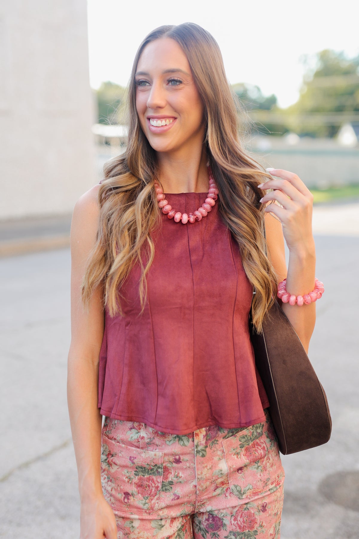 A woman wearing a sleeveless dusty rose top and floral pants, accessorized with a chunky pink beaded necklace and a brown shoulder bag, photographed outdoors.