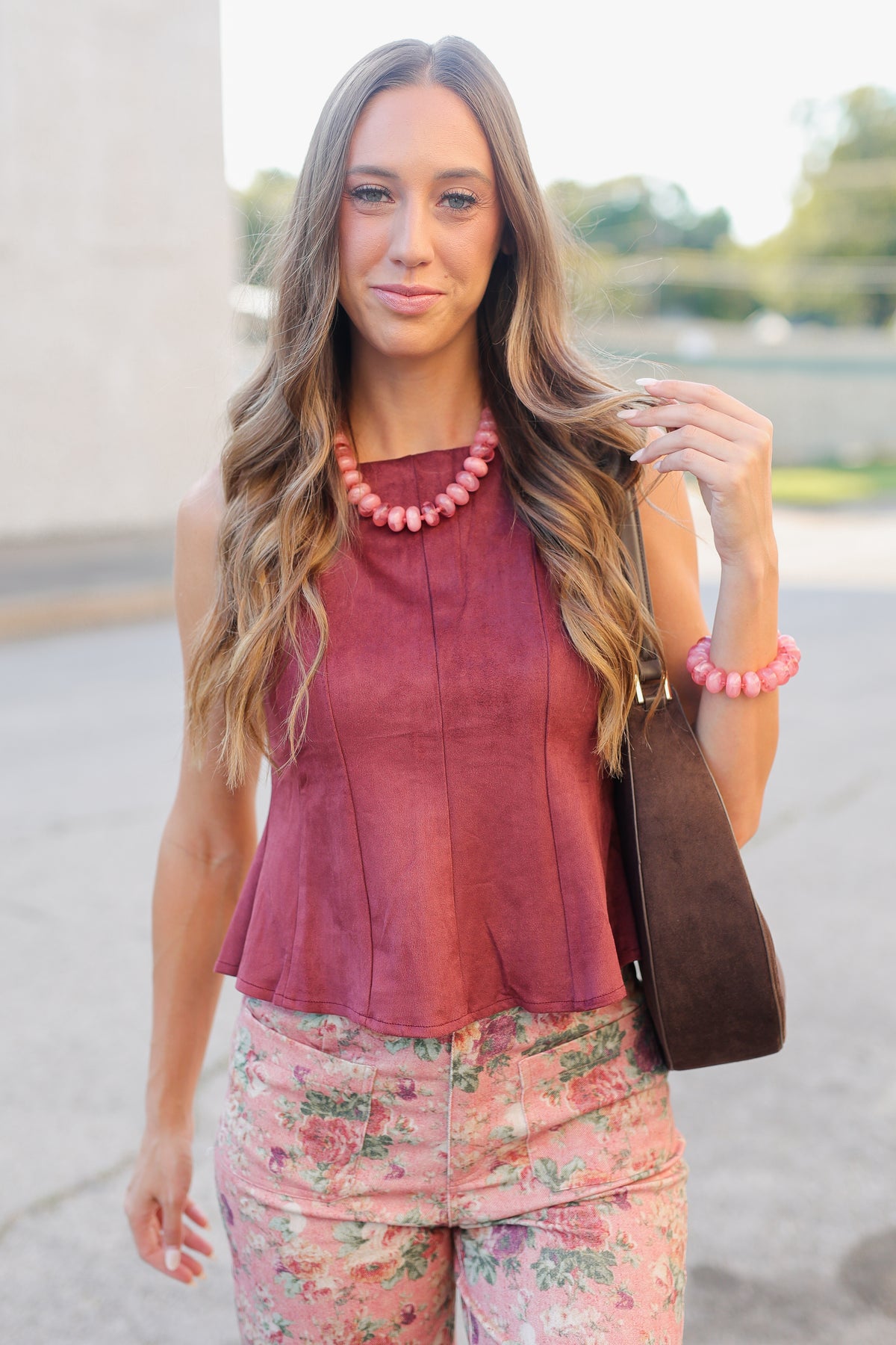 A woman wearing a sleeveless dusty rose top and floral pants, accessorized with a chunky pink beaded necklace and a brown shoulder bag, photographed outdoors.