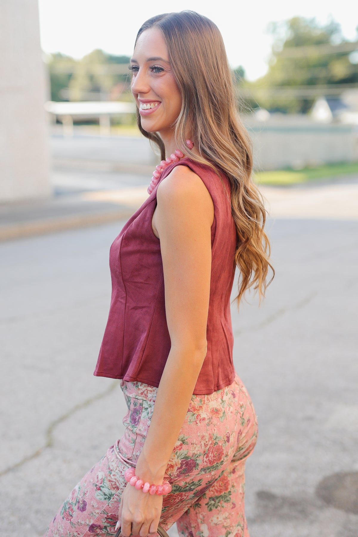 A woman wearing a sleeveless dusty rose top and pink floral pants, smiling and posing outdoors on a paved walkway.