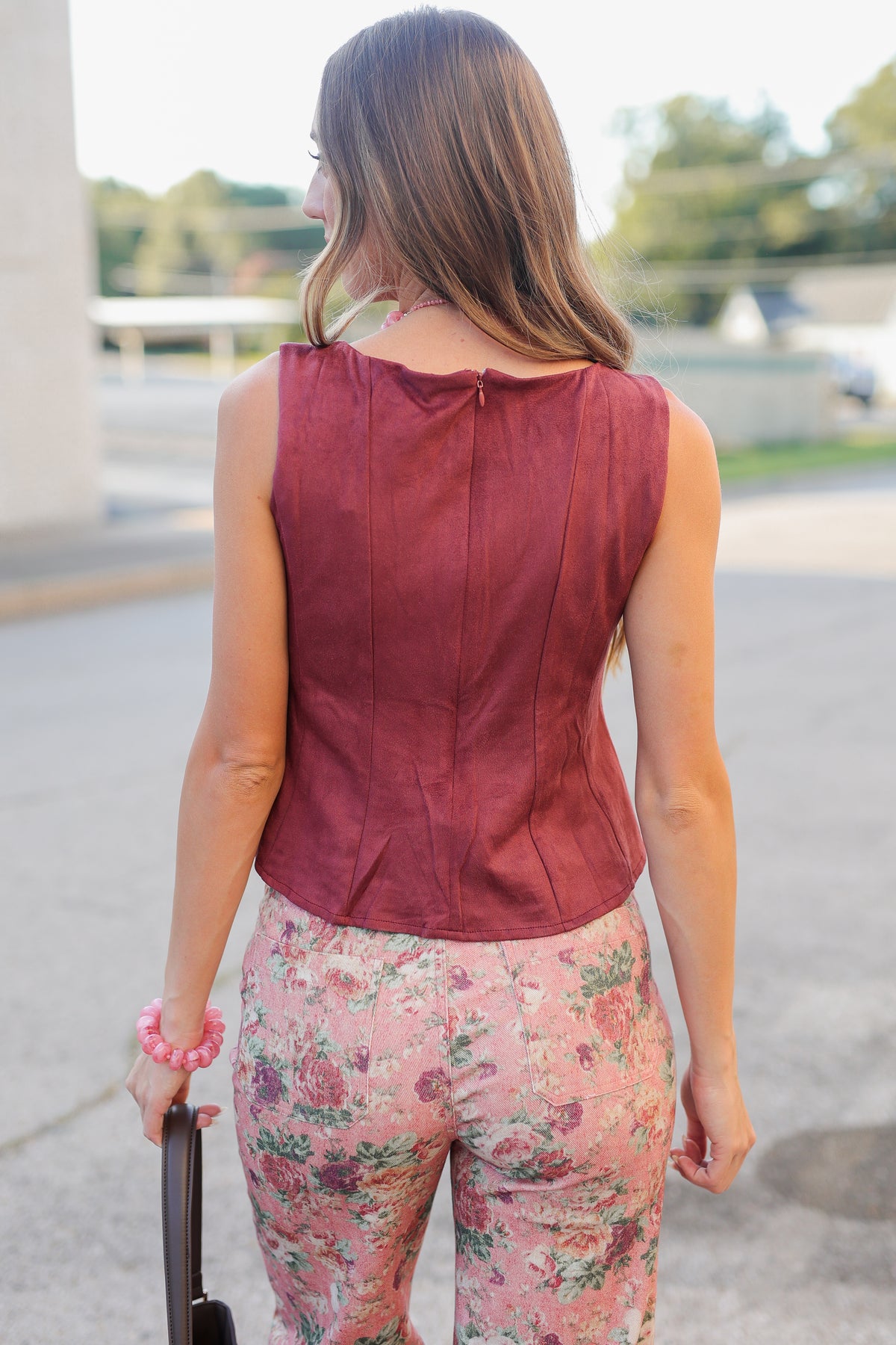 A woman wearing a sleeveless dusty rose top and floral pants, accessorized with a chunky pink beaded necklace and a brown shoulder bag, photographed outdoors.