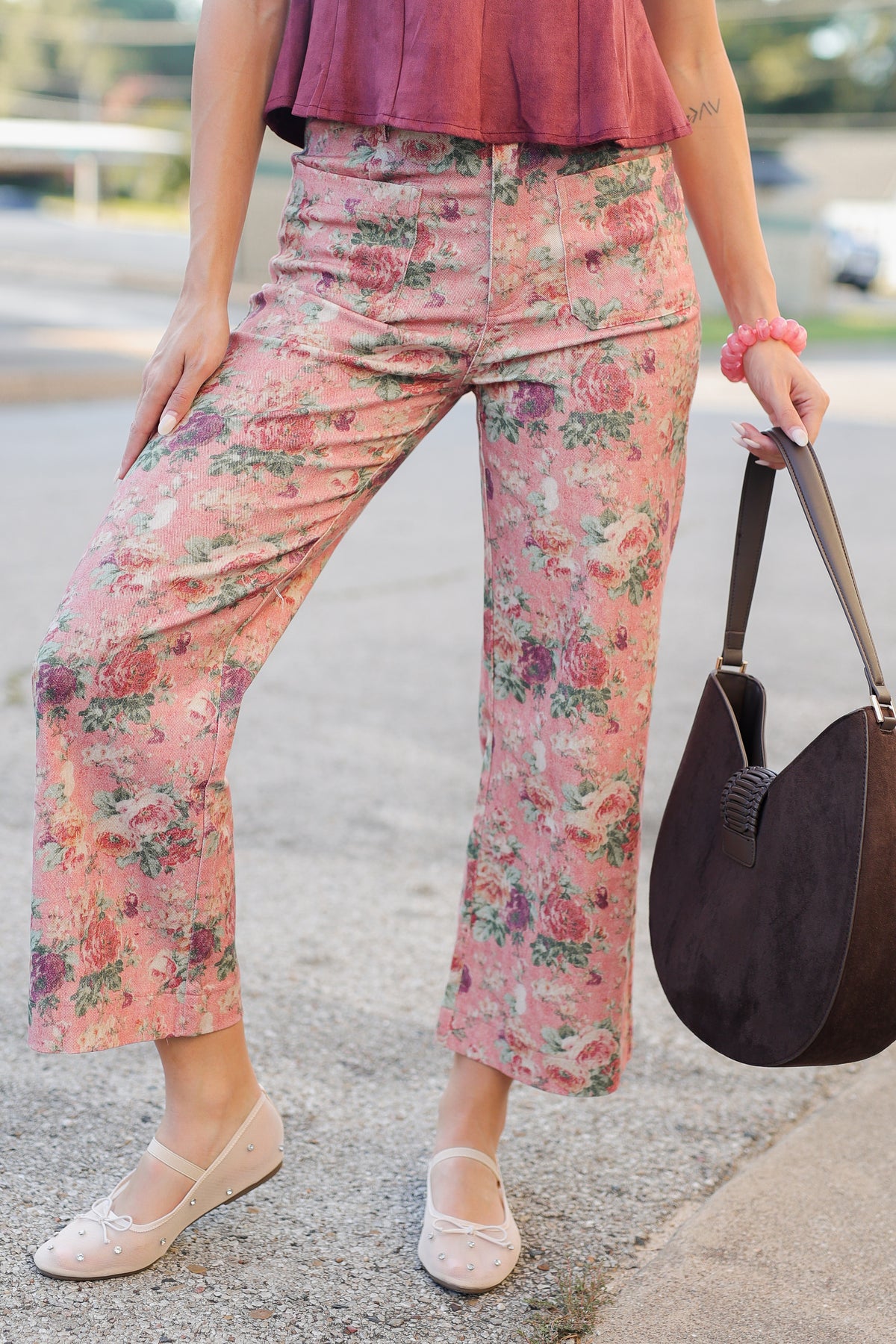 A person wearing pink floral cropped pants with a mauve top and beige flats, holding a dark brown shoulder bag outdoors on a paved surface.