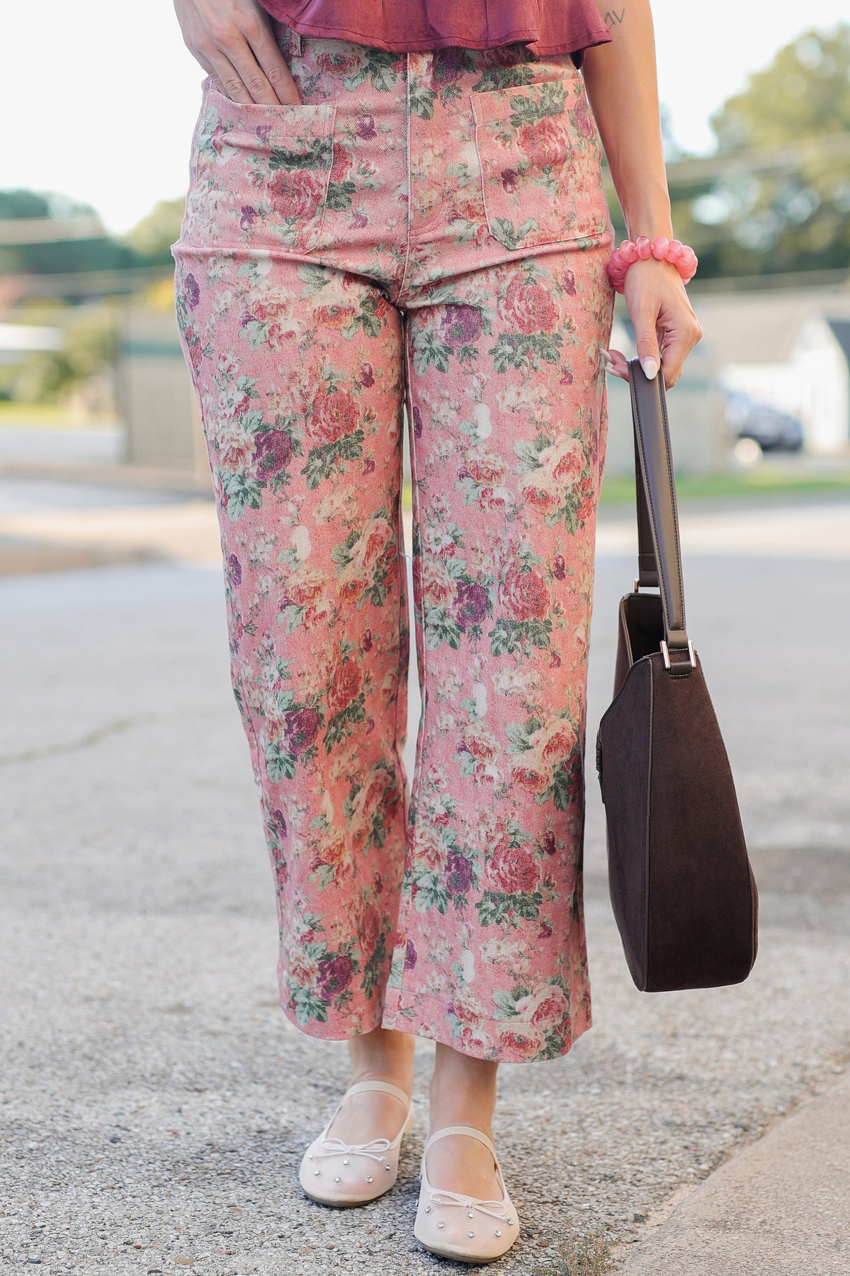 A person outdoors wearing pink floral cropped pants with a matching pink top, holding a dark brown shoulder bag, and wearing light pink flats.