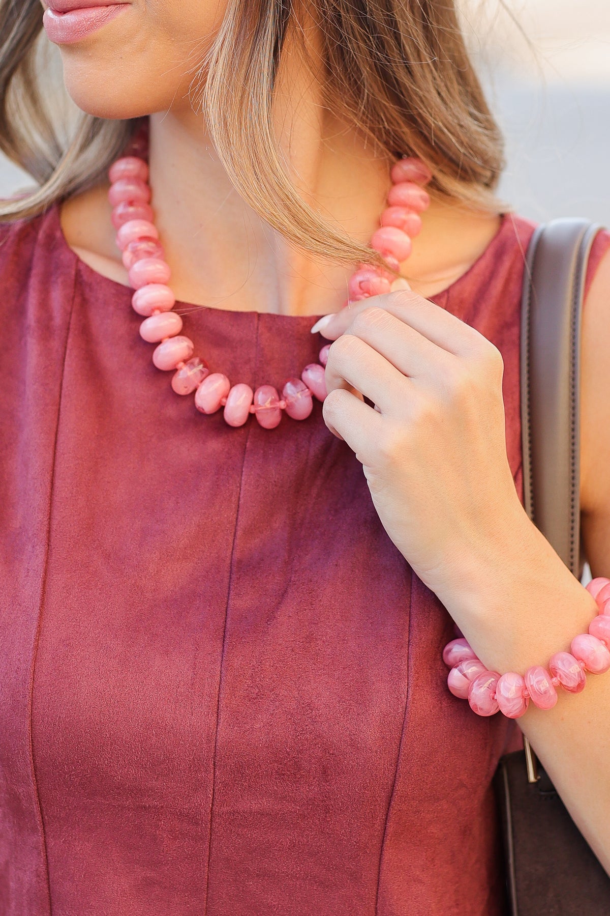 A close-up lifestyle shot of a woman wearing a chunky pink beaded necklace and matching pink beaded bracelet, paired with a dusty rose top.