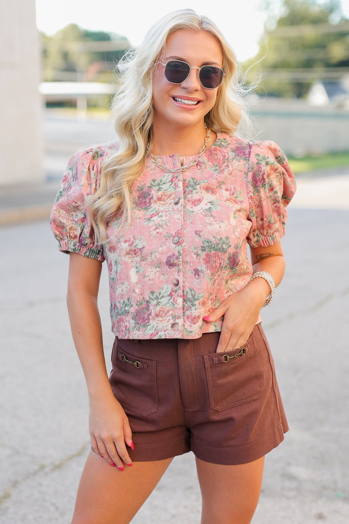 A woman wearing a pink floral puff-sleeve button-up top and brown shorts, standing outdoors on a paved walkway.