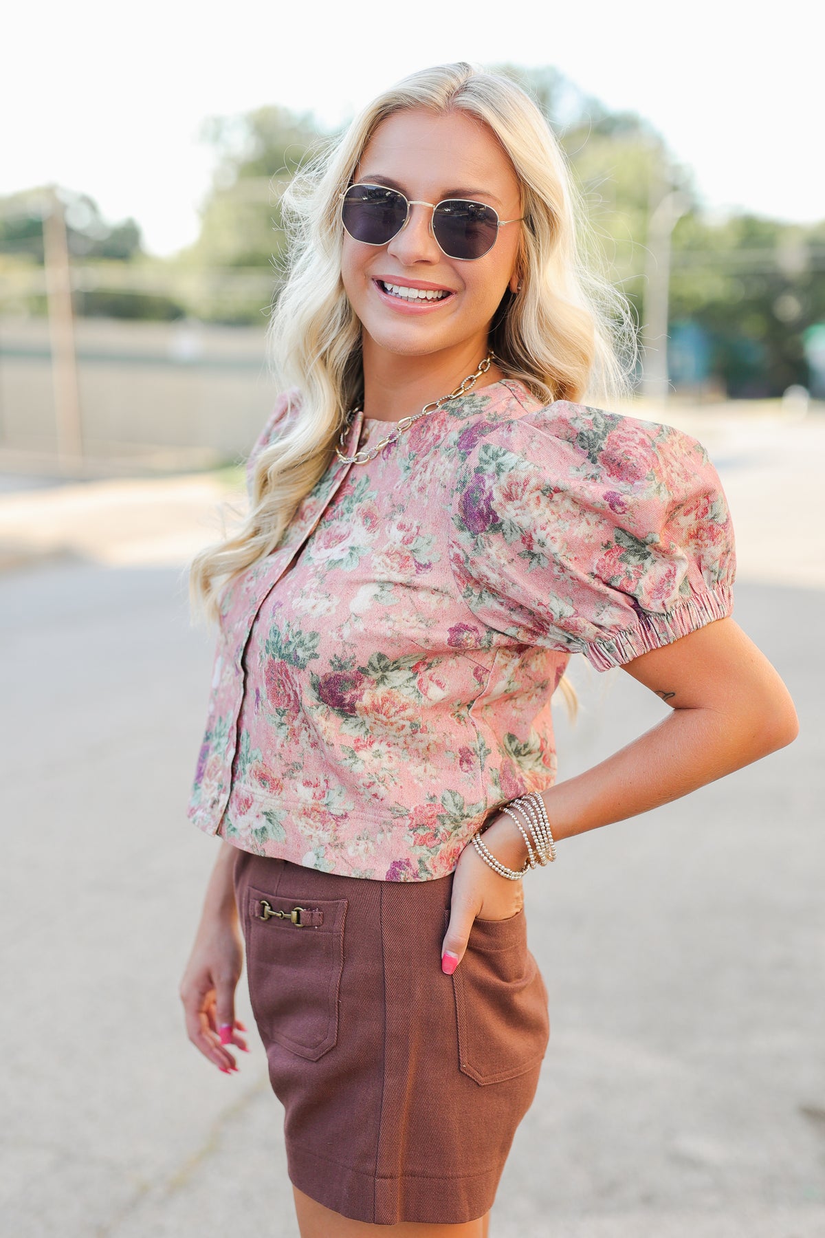 A woman wearing a pink floral puff-sleeve crop top and a brown mini skirt, standing outdoors on a paved surface with trees in the background.