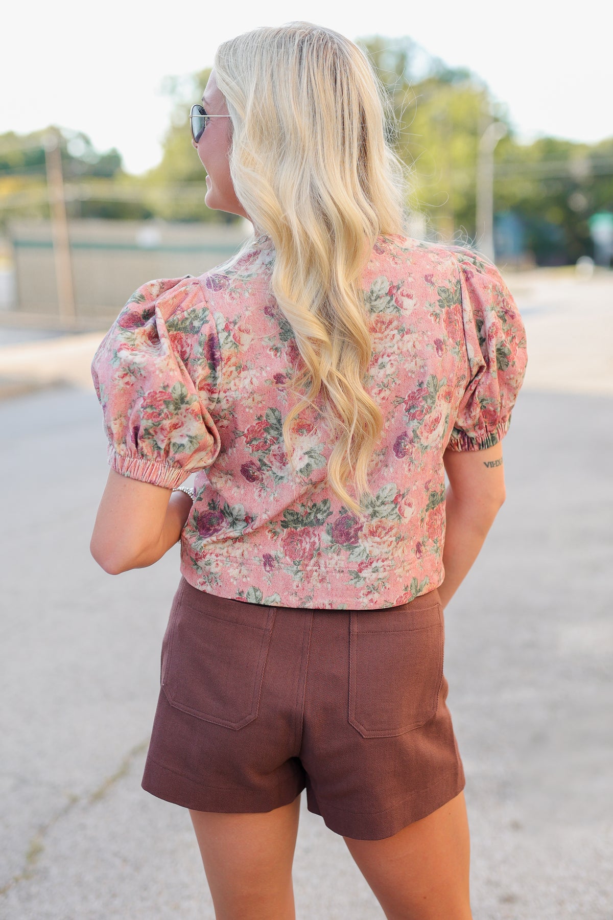 A woman viewed from behind wearing a pink floral puff-sleeve crop top and brown shorts, standing outdoors on a paved path.