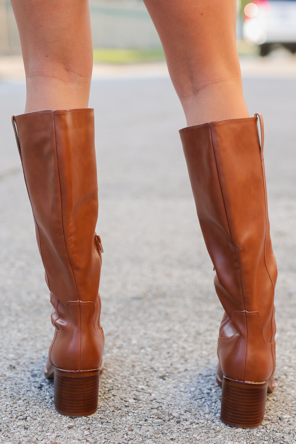 A close-up rear view of a person wearing knee-high brown leather boots with block heels, standing on a paved outdoor surface.