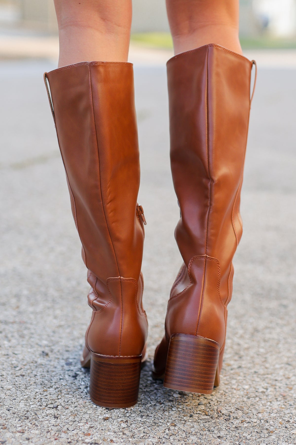 A pair of knee-high brown leather boots with stacked block heels, shown from the back on a person standing on asphalt.