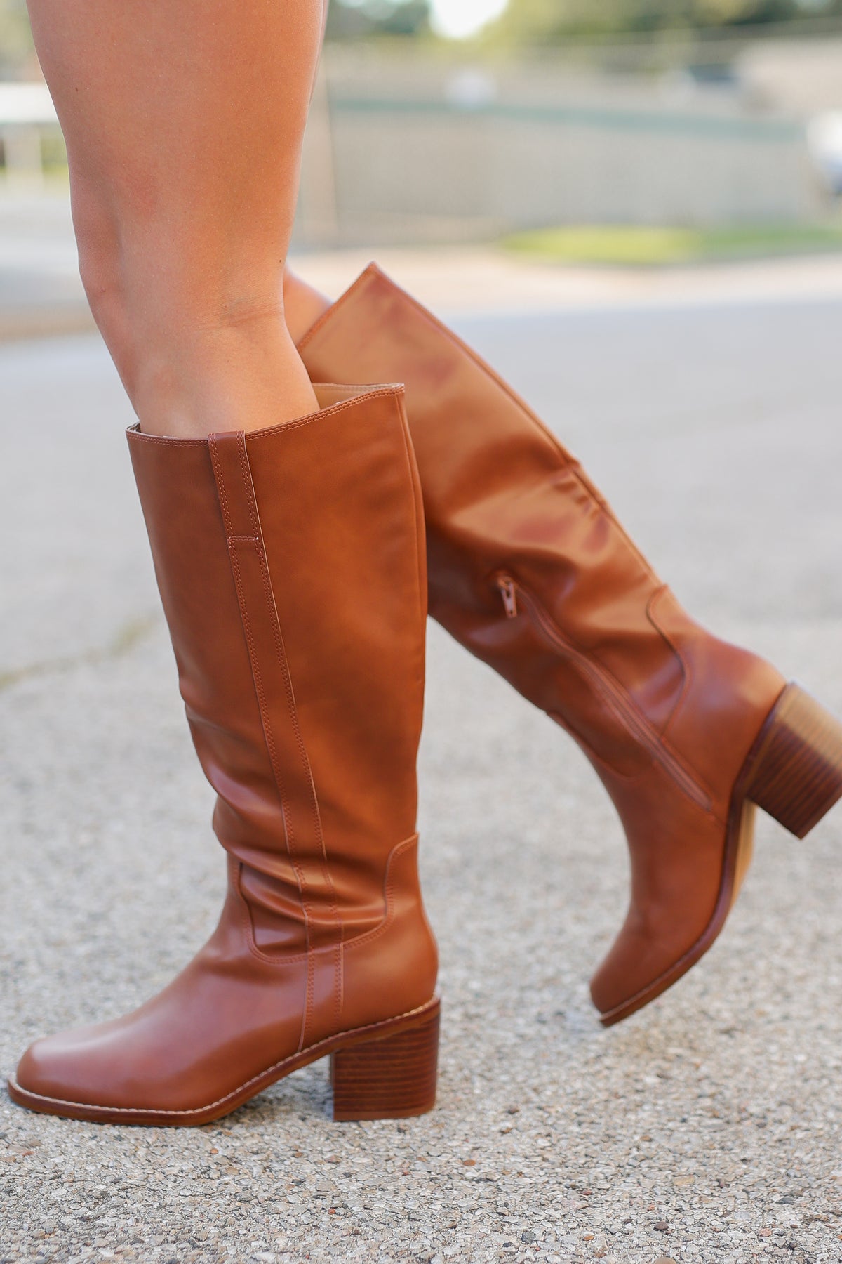 A pair of knee-high brown leather boots worn on a model's legs, standing on a paved outdoor surface.