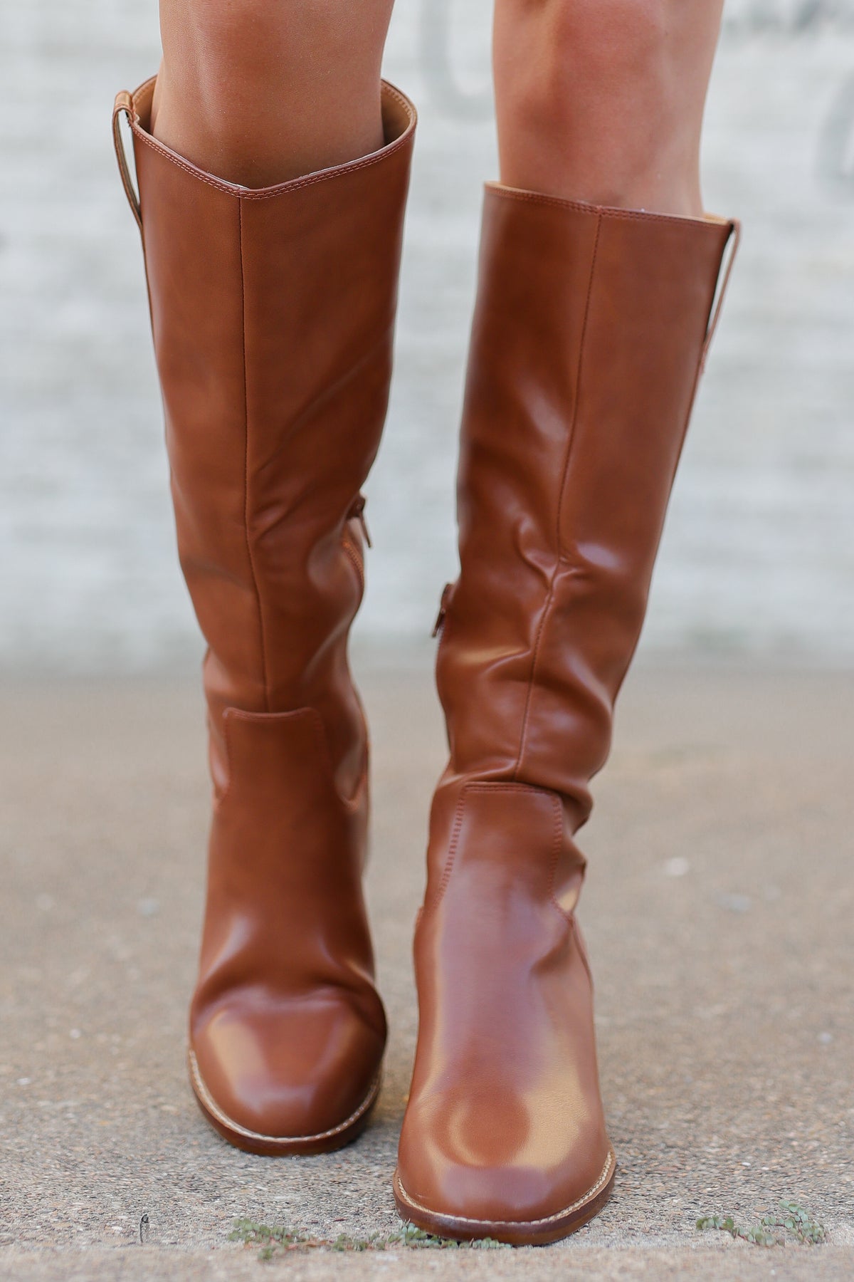A pair of knee-high brown leather boots with a slouchy, gathered design, shown worn on a person's legs against a concrete background.