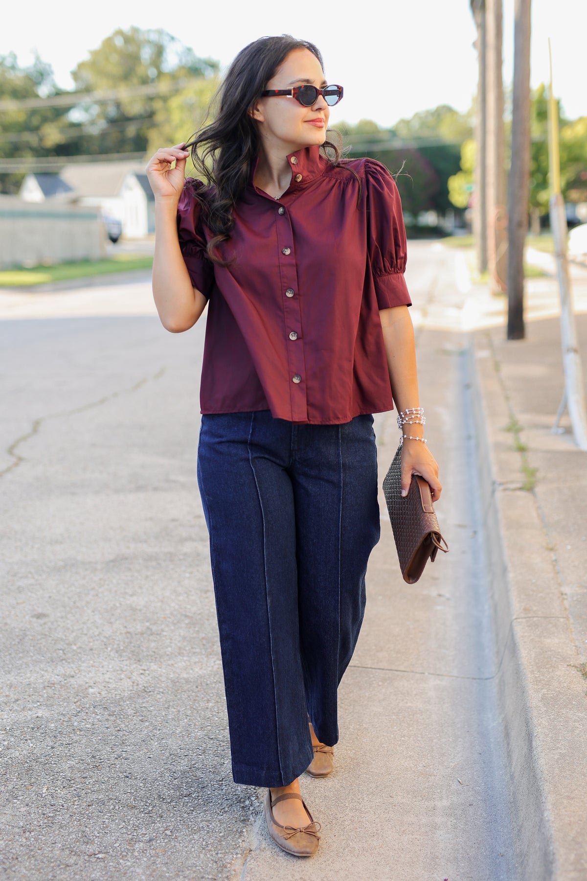 A woman walking outdoors on a sidewalk wearing a burgundy short-sleeve button-up blouse with puffed shoulders, dark wide-leg jeans, brown flats, sunglasses, and holding a brown clutch.