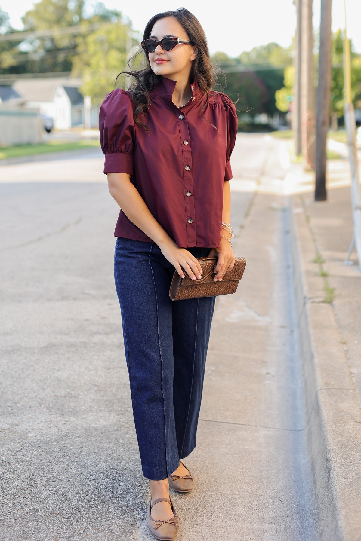 A woman walking outdoors on a sunny day, wearing a burgundy short-sleeve button-up blouse with puffed shoulders, dark blue wide-leg jeans, and tan flat sandals, while holding a brown leather clutch.