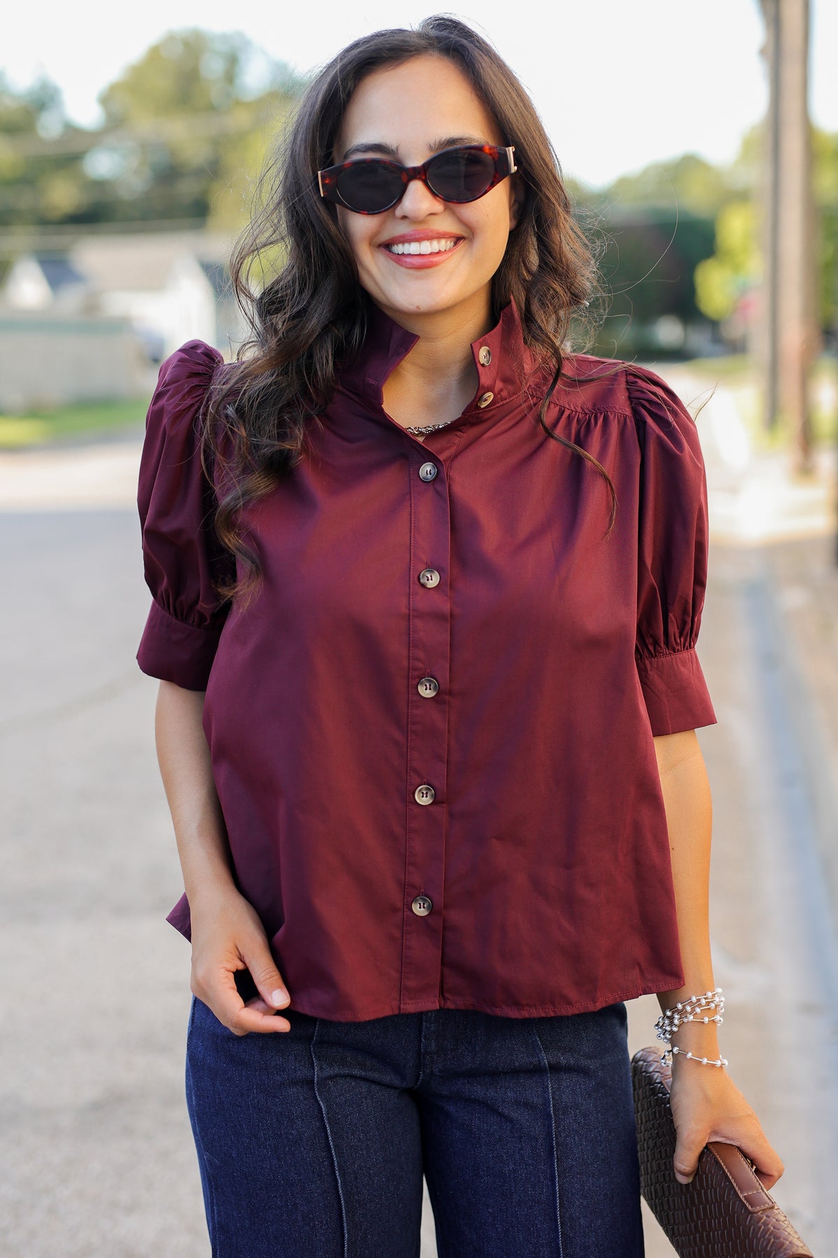 A woman wearing a burgundy short-sleeve blouse with puff sleeves and a stand collar, paired with dark blue jeans and sunglasses, standing outdoors on a sunny day.