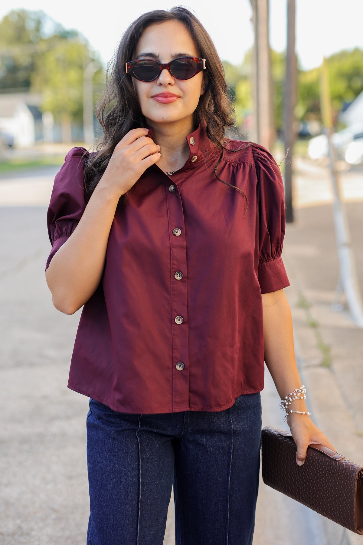 A woman wearing a burgundy short-sleeve button-up blouse with puff sleeves and dark blue jeans, standing outdoors on a sidewalk.