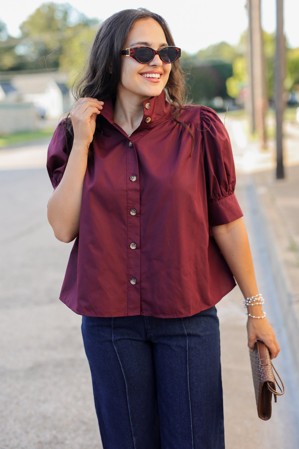 A woman wearing a burgundy short-sleeve button-up blouse with puff sleeves and dark blue jeans, standing outdoors on a sunny day.
