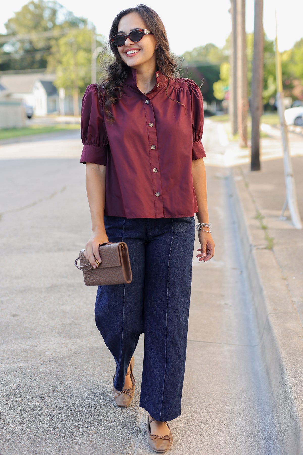 A woman walking outdoors on a sunny street wearing a burgundy short-sleeve blouse with puffed shoulders, dark wide-leg jeans, tan flats, and holding a brown clutch.