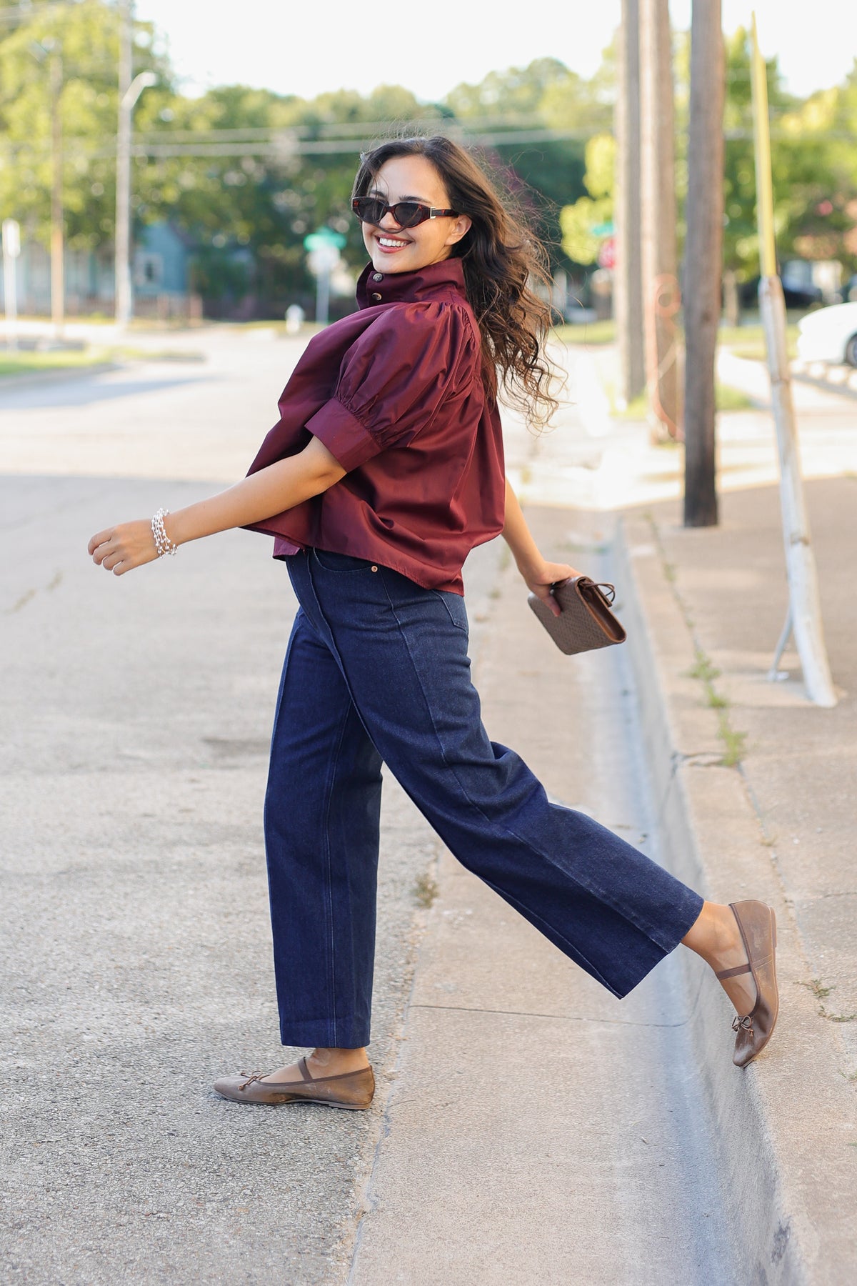 A woman walking outdoors on a sidewalk wearing a burgundy short-sleeve blouse, navy wide-leg pants, brown flat shoes, and sunglasses, holding a small brown clutch.