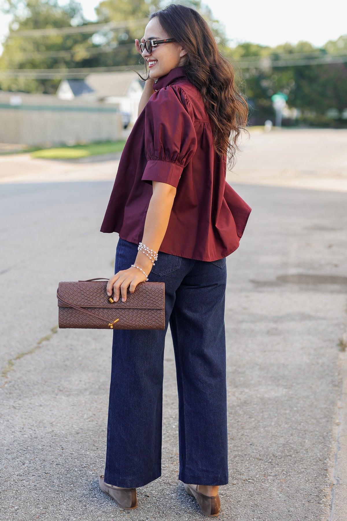 A woman wearing a burgundy short-sleeve puff-sleeve blouse, dark blue wide-leg jeans, and brown shoes, holding a brown quilted clutch bag, standing outdoors on a paved street.
