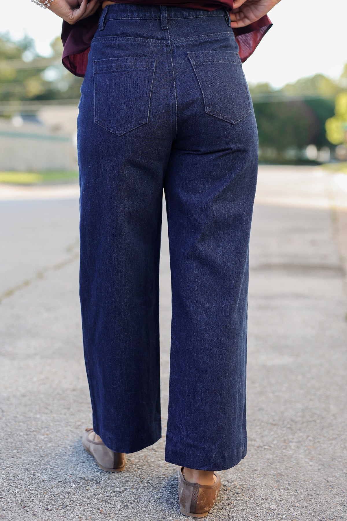 A person shown from the back wearing dark blue wide-leg cropped jeans, standing outdoors on a paved surface with trees in the background.