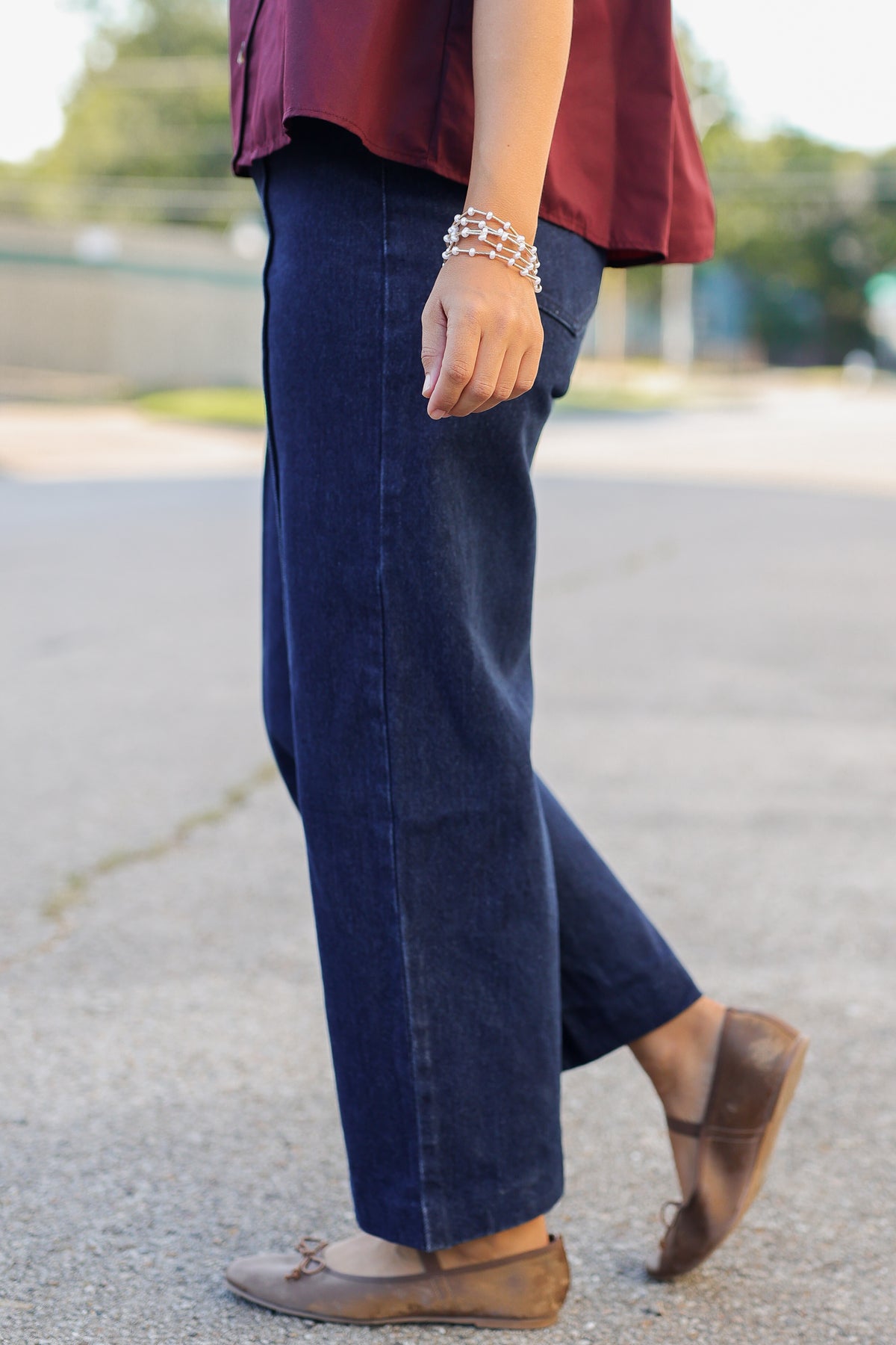 A person wearing dark blue wide-leg cropped jeans, a burgundy top, and brown flat shoes, standing outdoors on a paved surface.