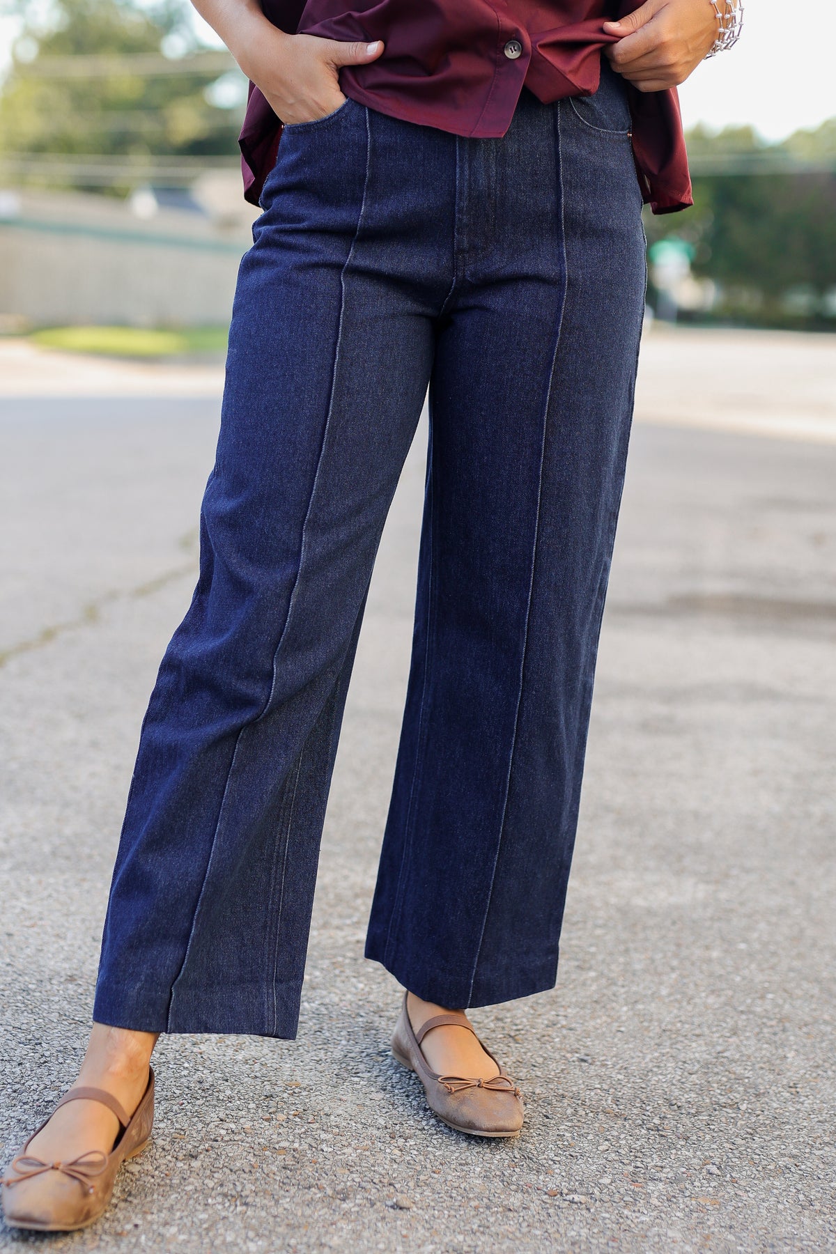 A person wearing dark blue wide-leg denim pants with a burgundy top and tan flat shoes, standing outdoors on a paved surface.