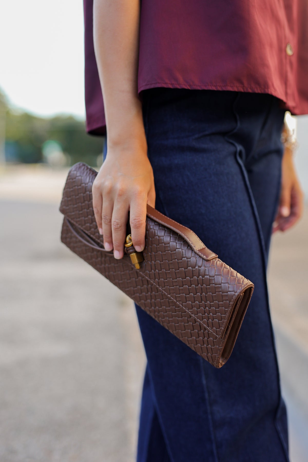 A person wearing a burgundy top and dark blue jeans holds a brown woven leather clutch bag with a gold-tone clasp outdoors.